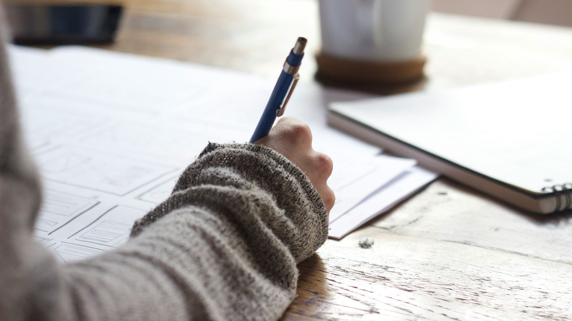 person writing on brown wooden table near white ceramic mug