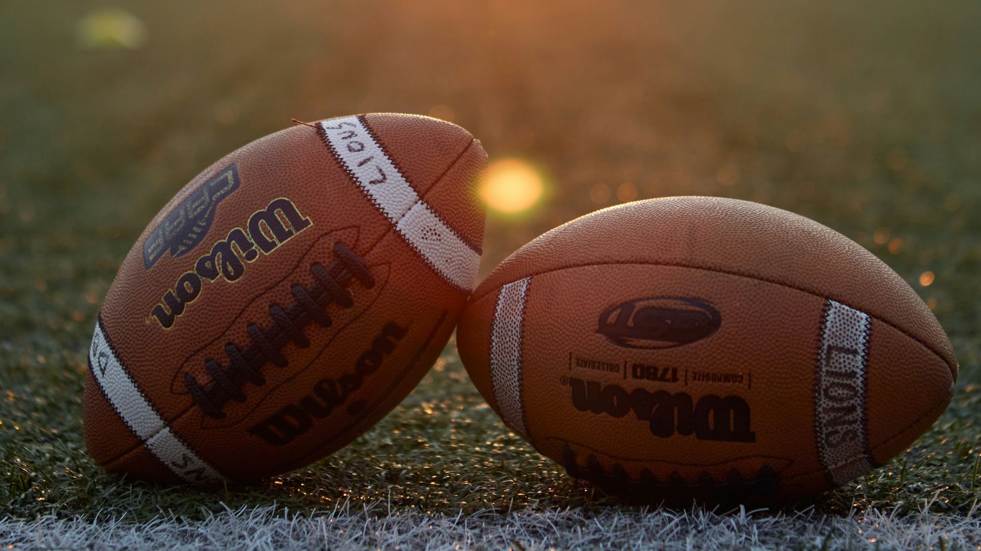 Two footballs sitting on a football field at sunset