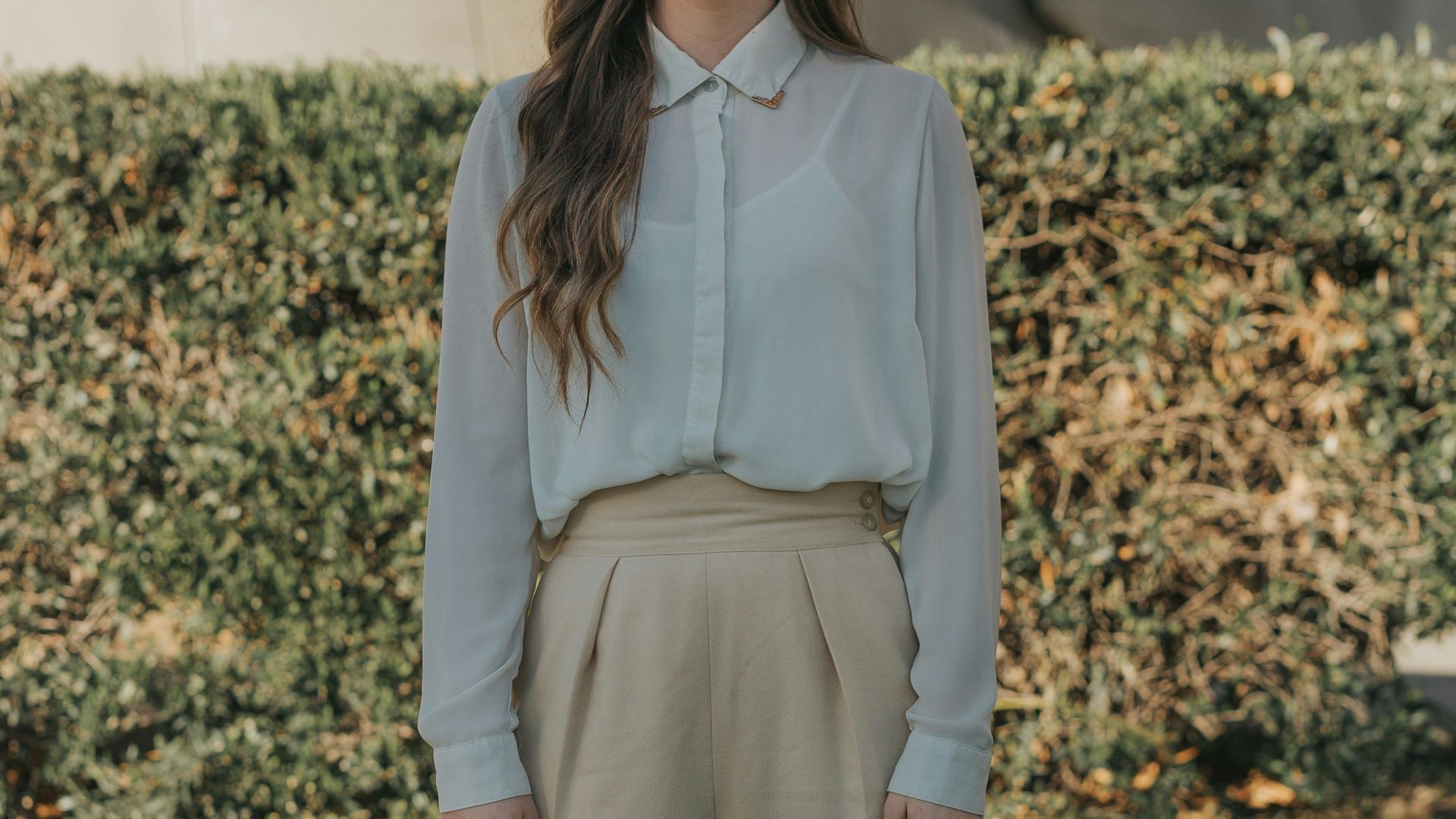 woman wearing gray button-up long-sleeved shirt and brown pants standing in front of bushes