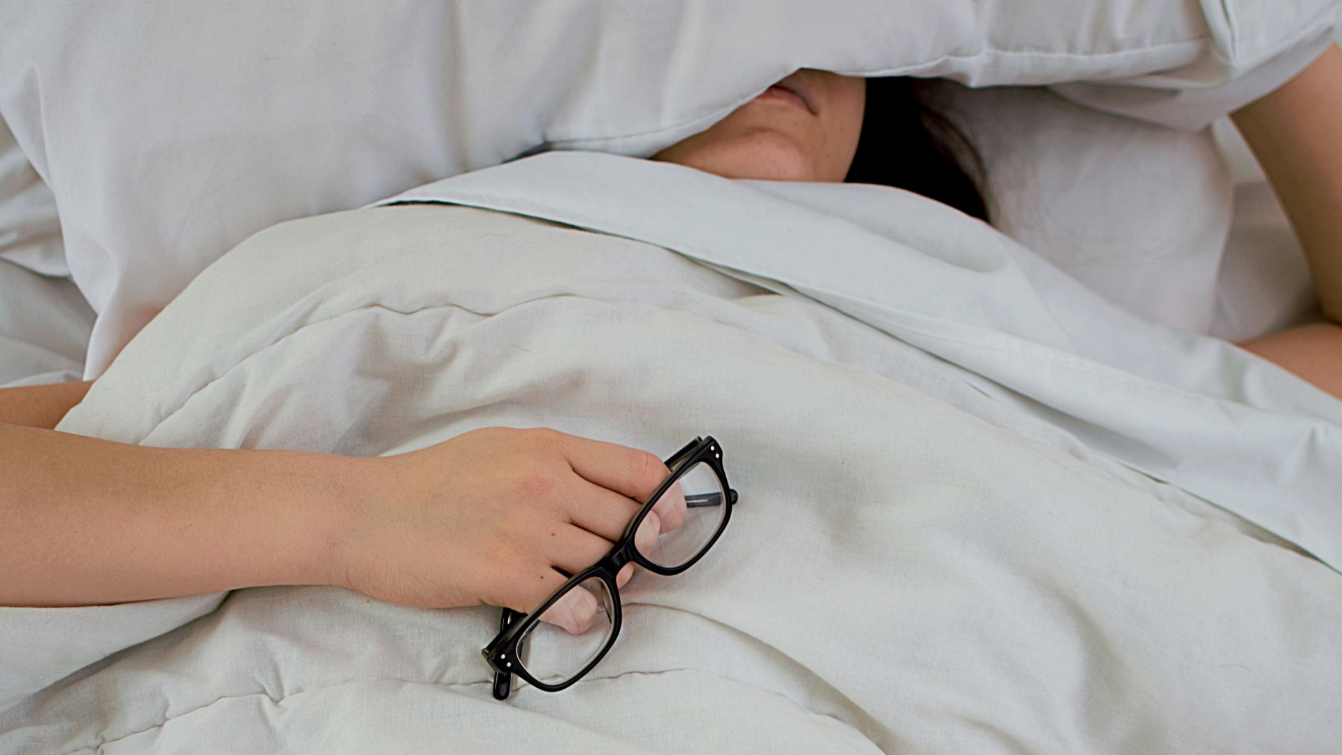 person lying on bed while covering face with pillow and holding eyeglasses