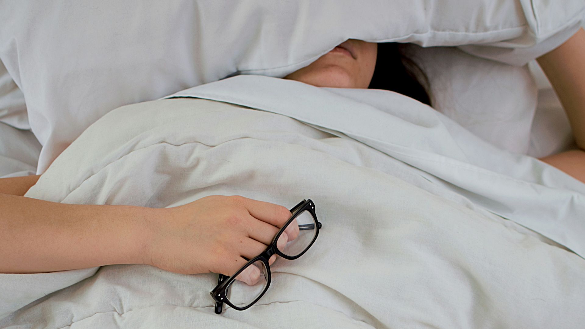 person lying on bed while covering face with pillow and holding eyeglasses