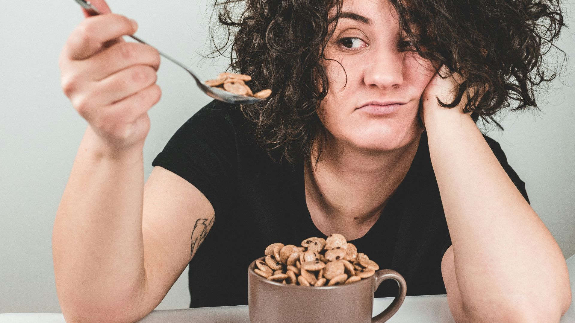 woman with messy hair wearing black crew-neck t-shirt holding spoon with cereals on top