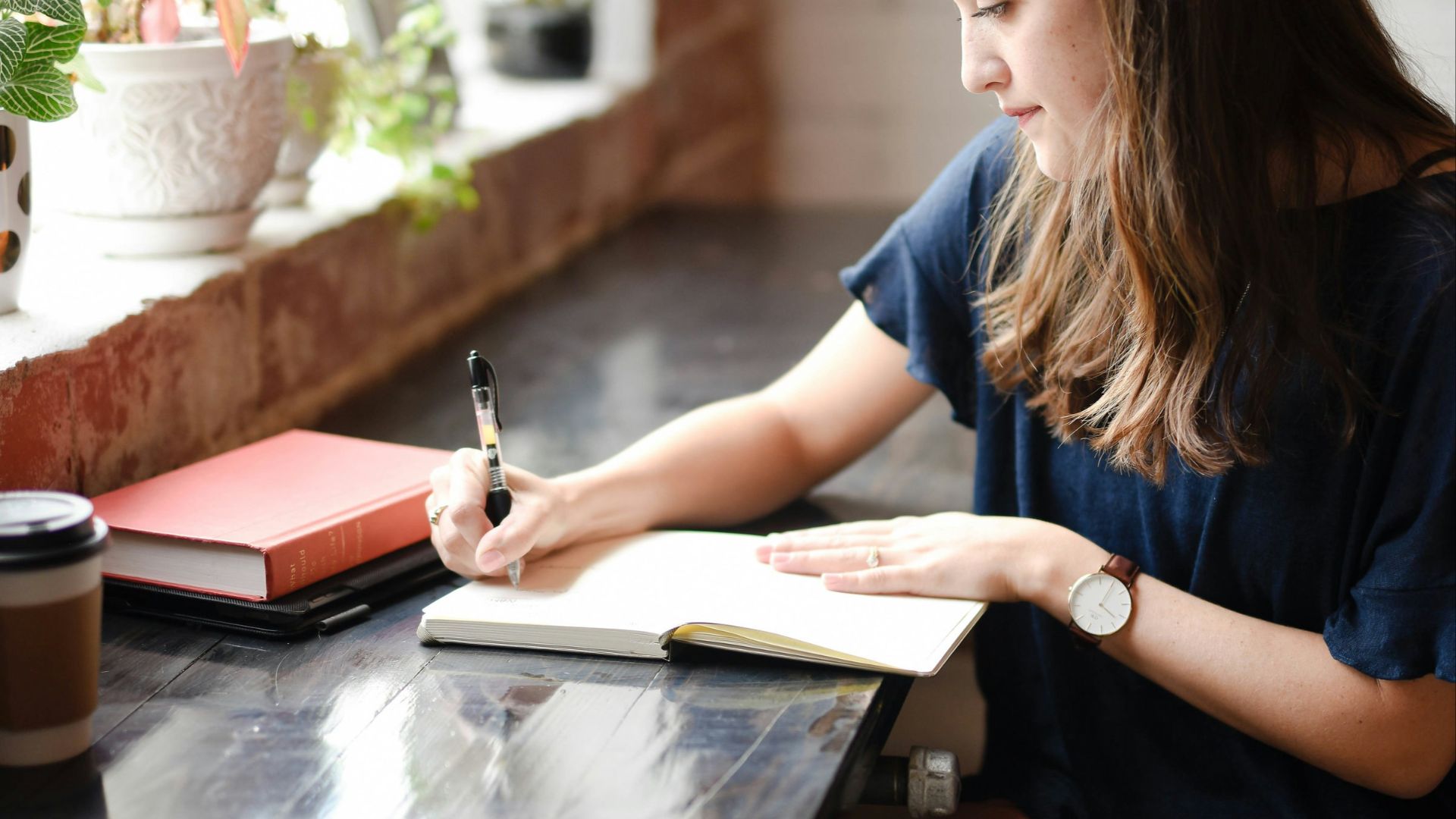 woman sitting in front of black table writing on white book near window
