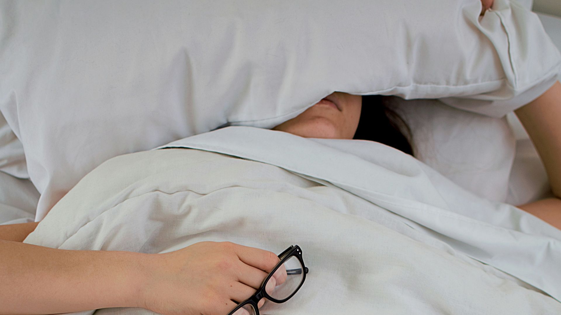 person lying on bed while covering face with pillow and holding eyeglasses