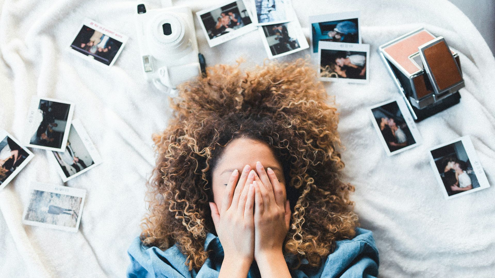 woman lying on bed covering her face surrounded by photos and white camera