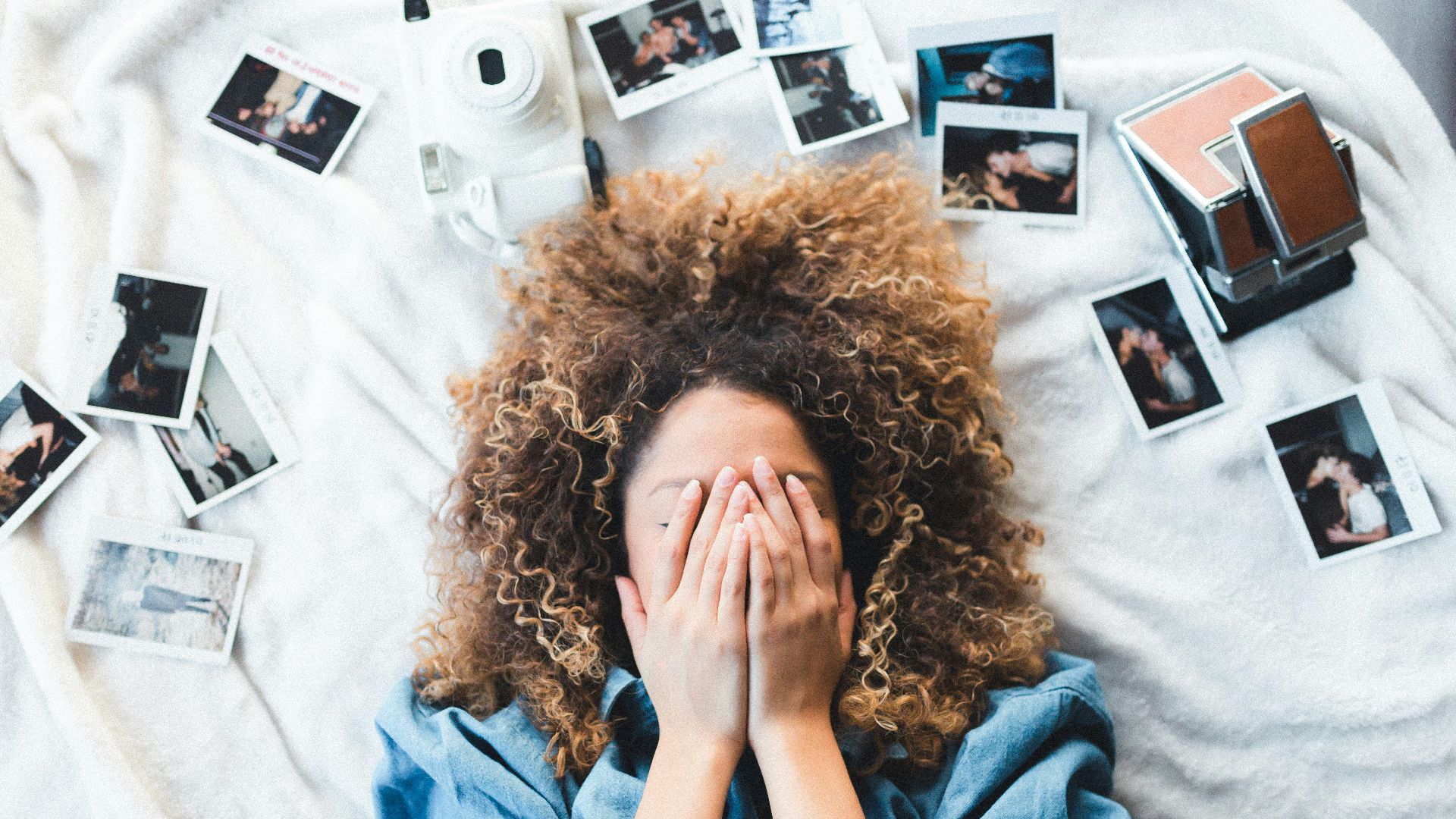 woman lying on bed covering her face surrounded by photos and white camera