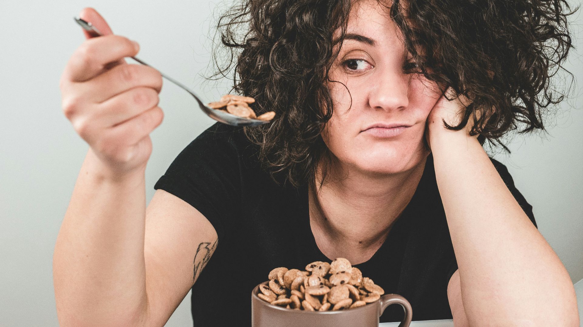 woman with messy hair wearing black crew-neck t-shirt holding spoon with cereals on top