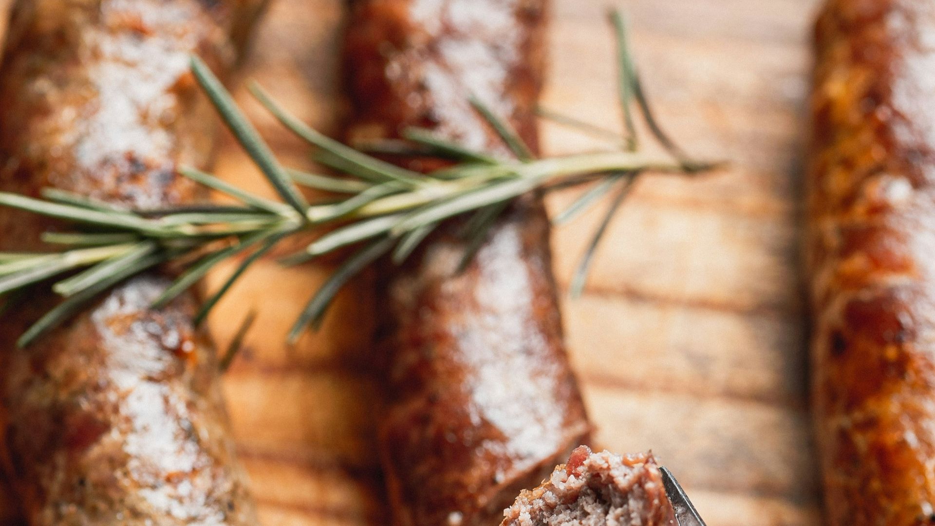 Three browned sausages on a wooden board with rosemary.