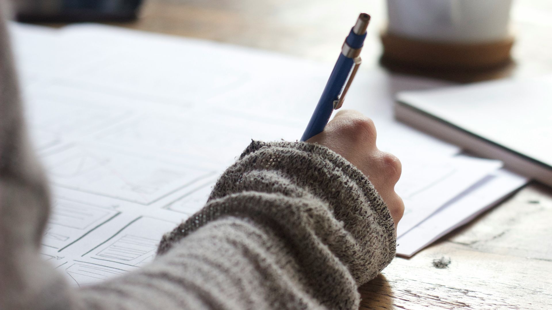 person writing on brown wooden table near white ceramic mug