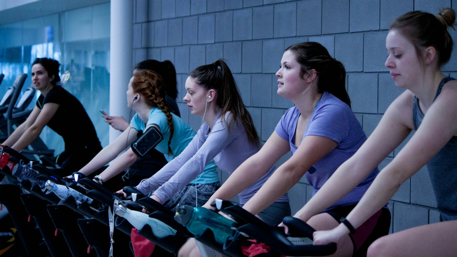 women taking exercise on black stationary bikes in front of gray concrete wall