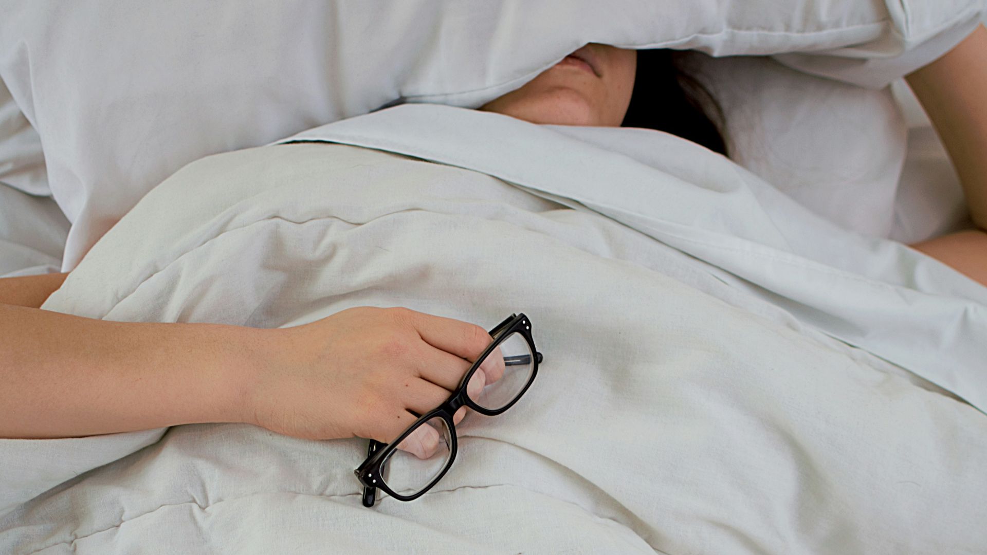person lying on bed while covering face with pillow and holding eyeglasses
