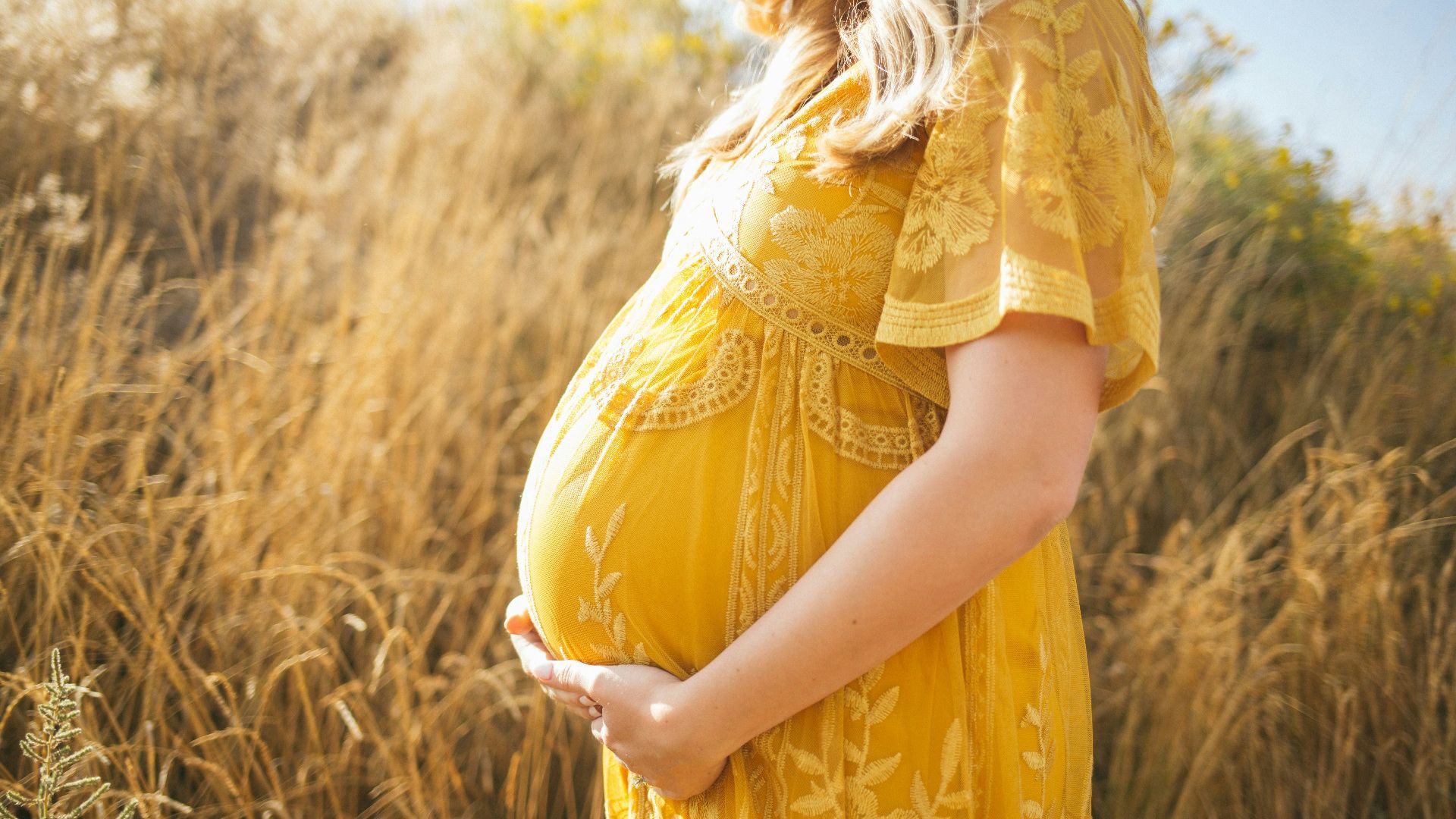 pregnant woman wearing yellow floral dress standing while touching her tummy and facing her right side near brown field during daytime