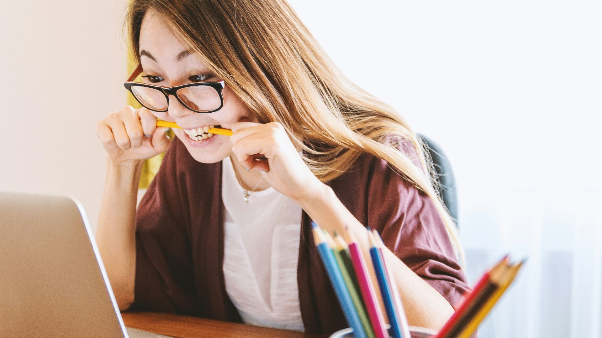woman biting pencil while sitting on chair in front of computer during daytime