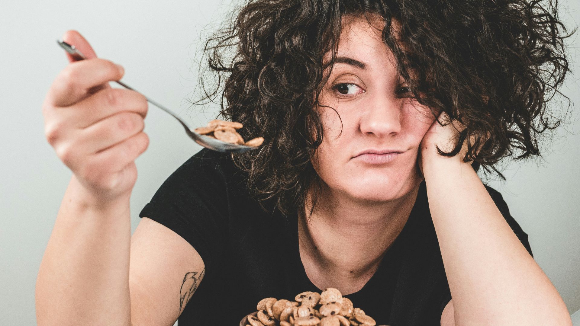woman with messy hair wearing black crew-neck t-shirt holding spoon with cereals on top
