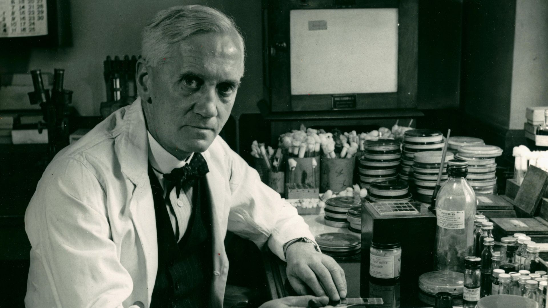 File:Alexander Fleming at his desk in a white lab coat with penicillin mold in petrie dishes around him.jpg
