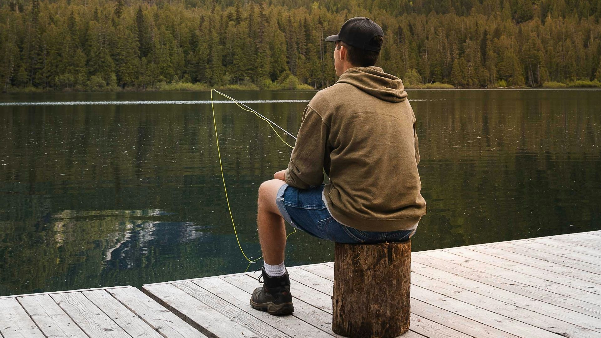 man in brown t-shirt and blue shorts standing on dock looking at lake during daytime