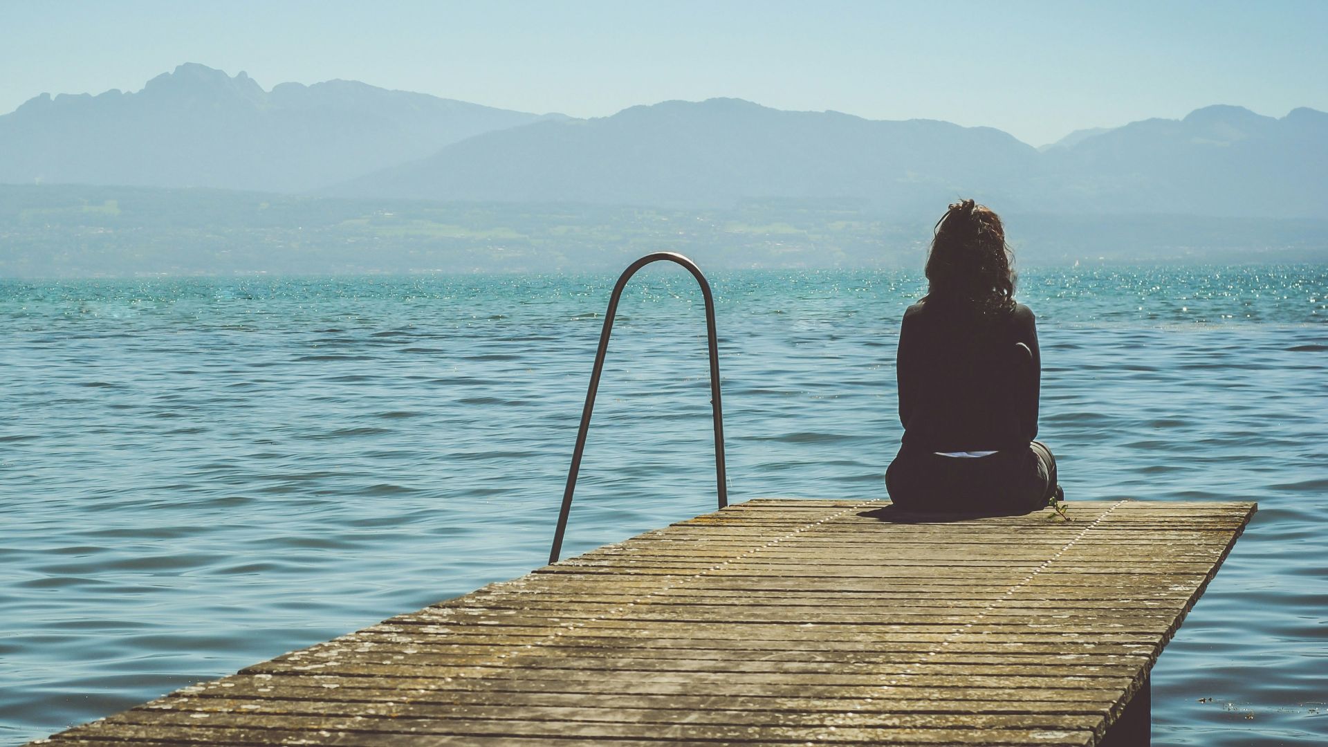 a woman sits on the end of a dock during daytime staring across a lake