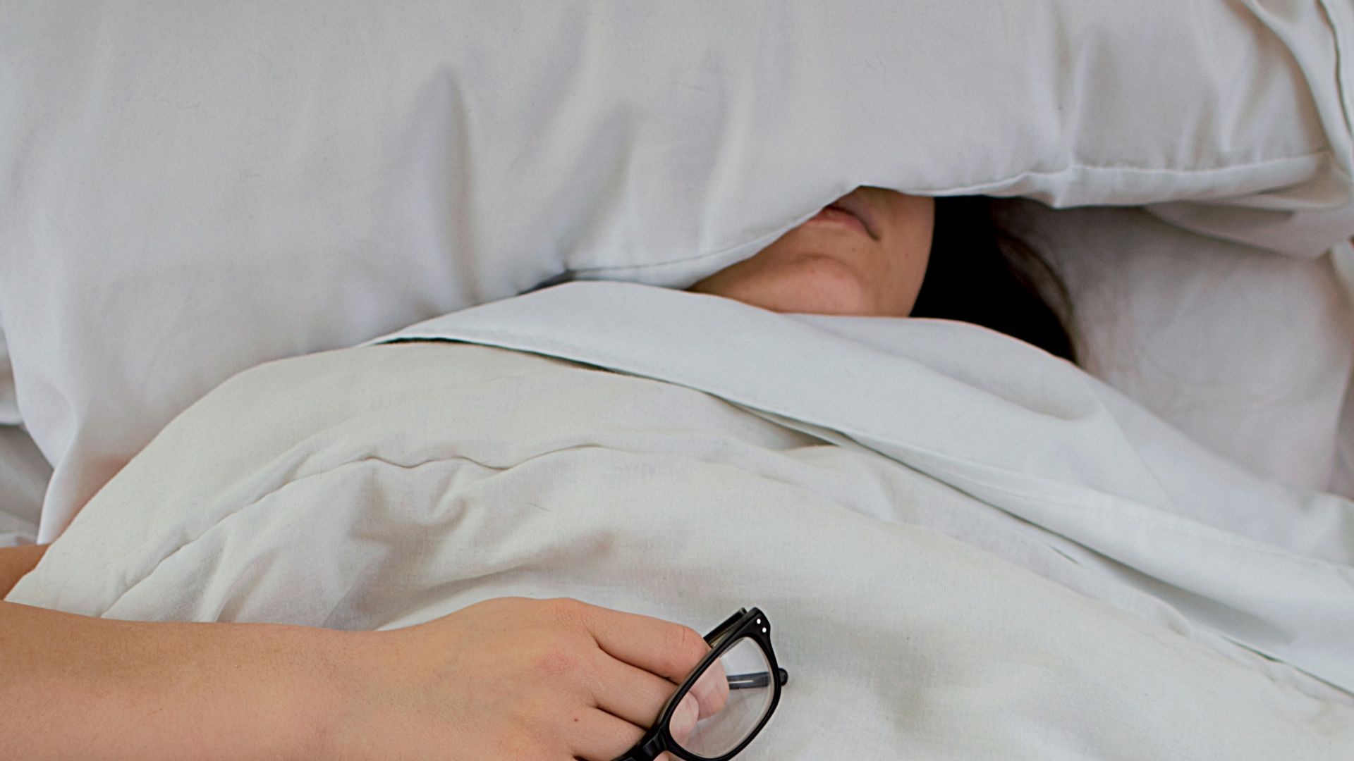person lying on bed while covering face with pillow and holding eyeglasses