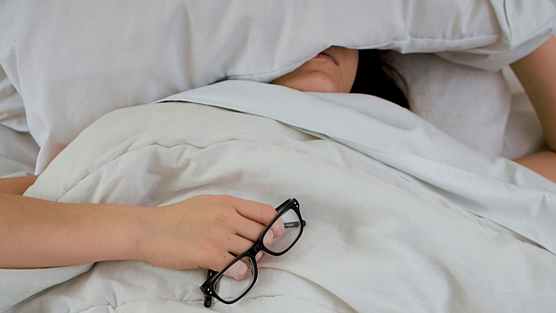 person lying on bed while covering face with pillow and holding eyeglasses