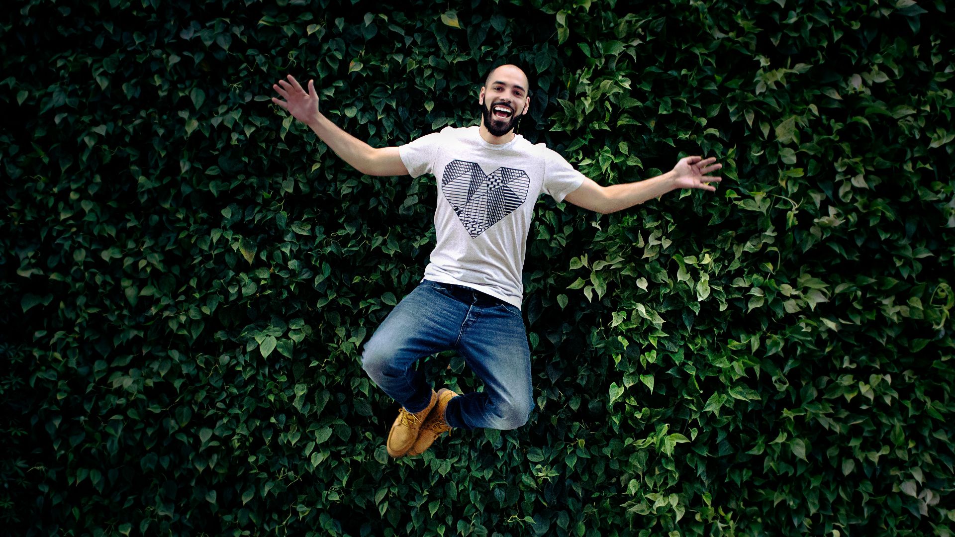 man wearing t-shirt and jeans jumpshot in front of a green hedge