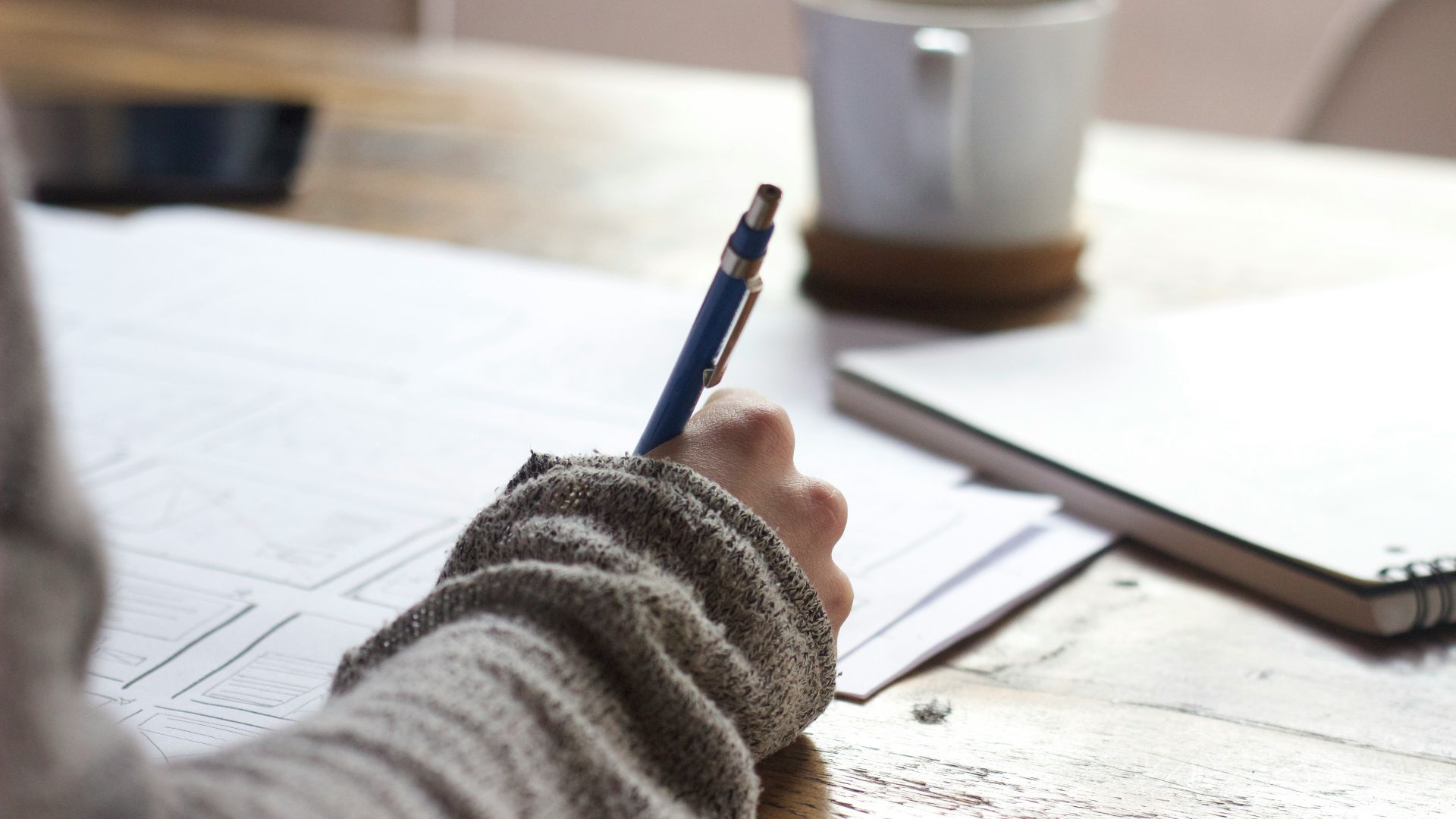 person writing on brown wooden table near white ceramic mug