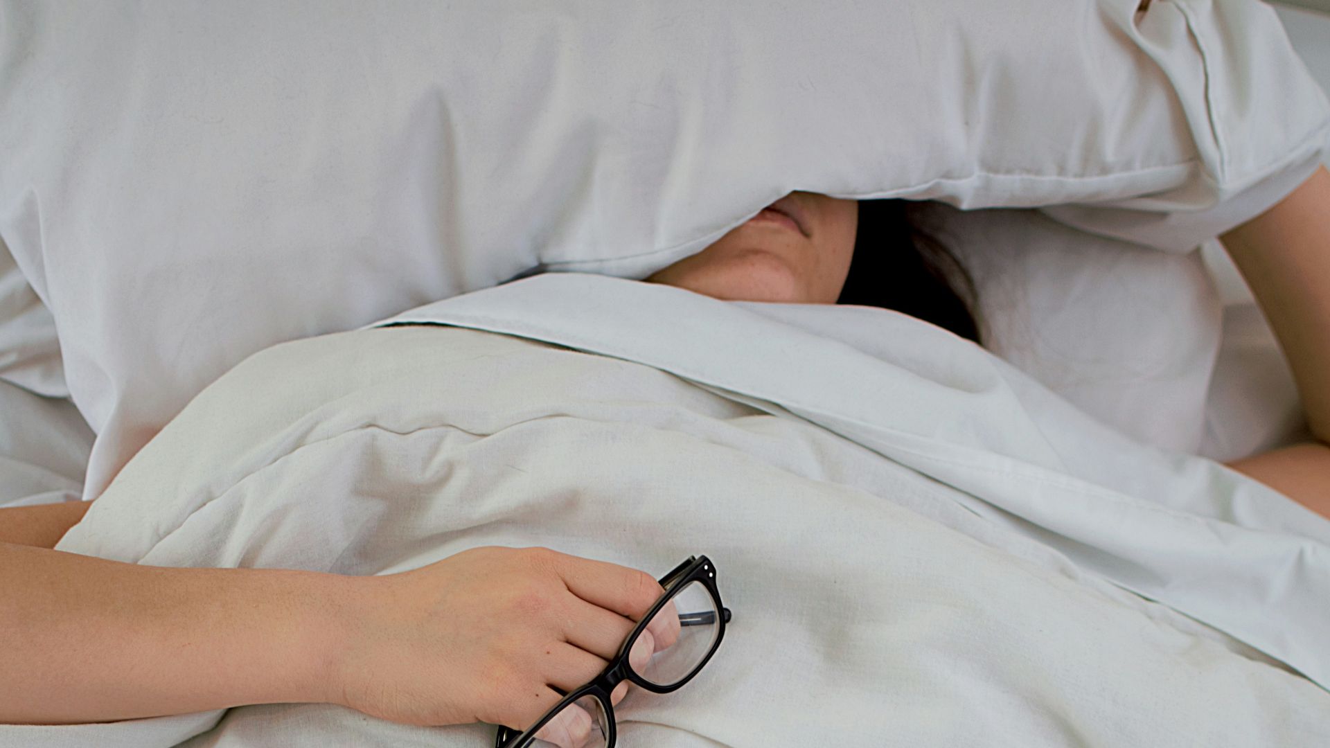 person lying on bed while covering face with pillow and holding eyeglasses