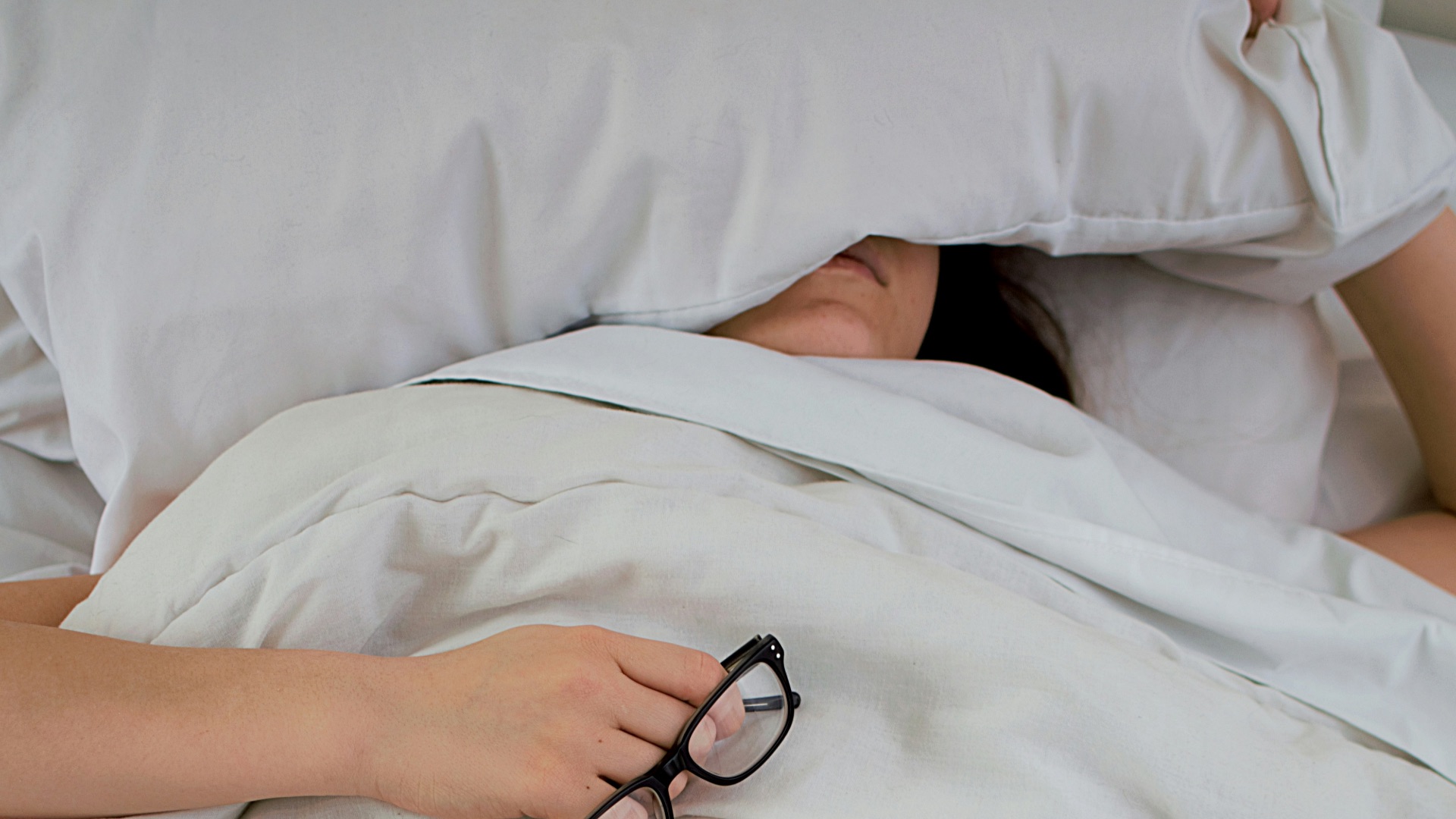 person lying on bed while covering face with pillow and holding eyeglasses