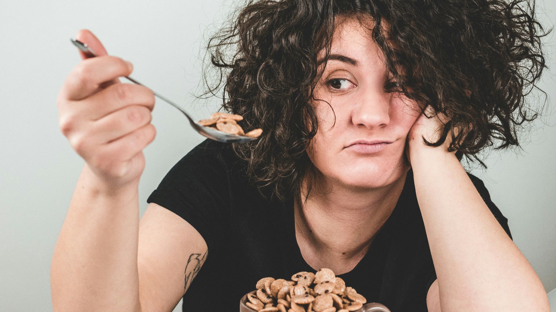 woman with messy hair wearing black crew-neck t-shirt holding spoon with cereals on top