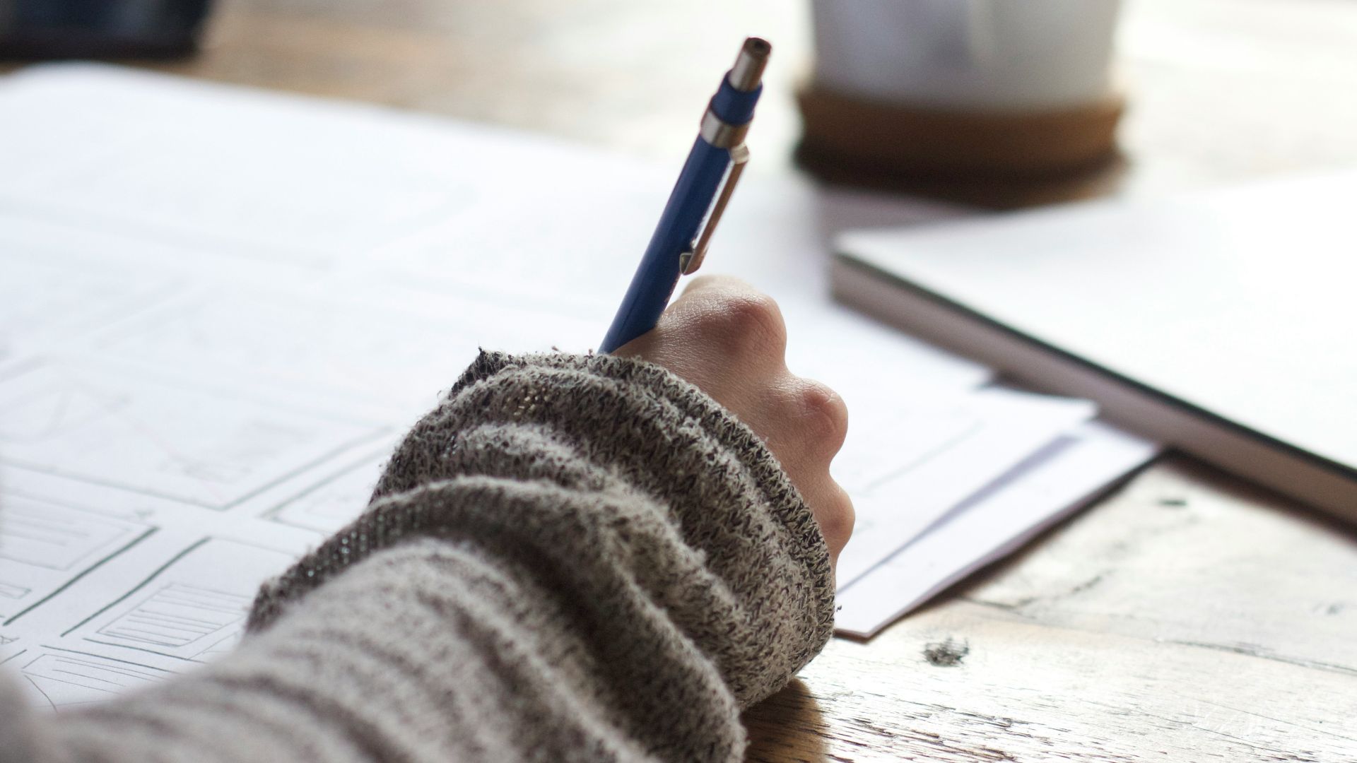 person writing on brown wooden table near white ceramic mug