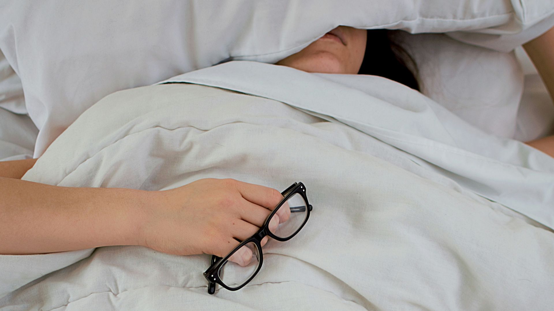 person lying on bed while covering face with pillow and holding eyeglasses