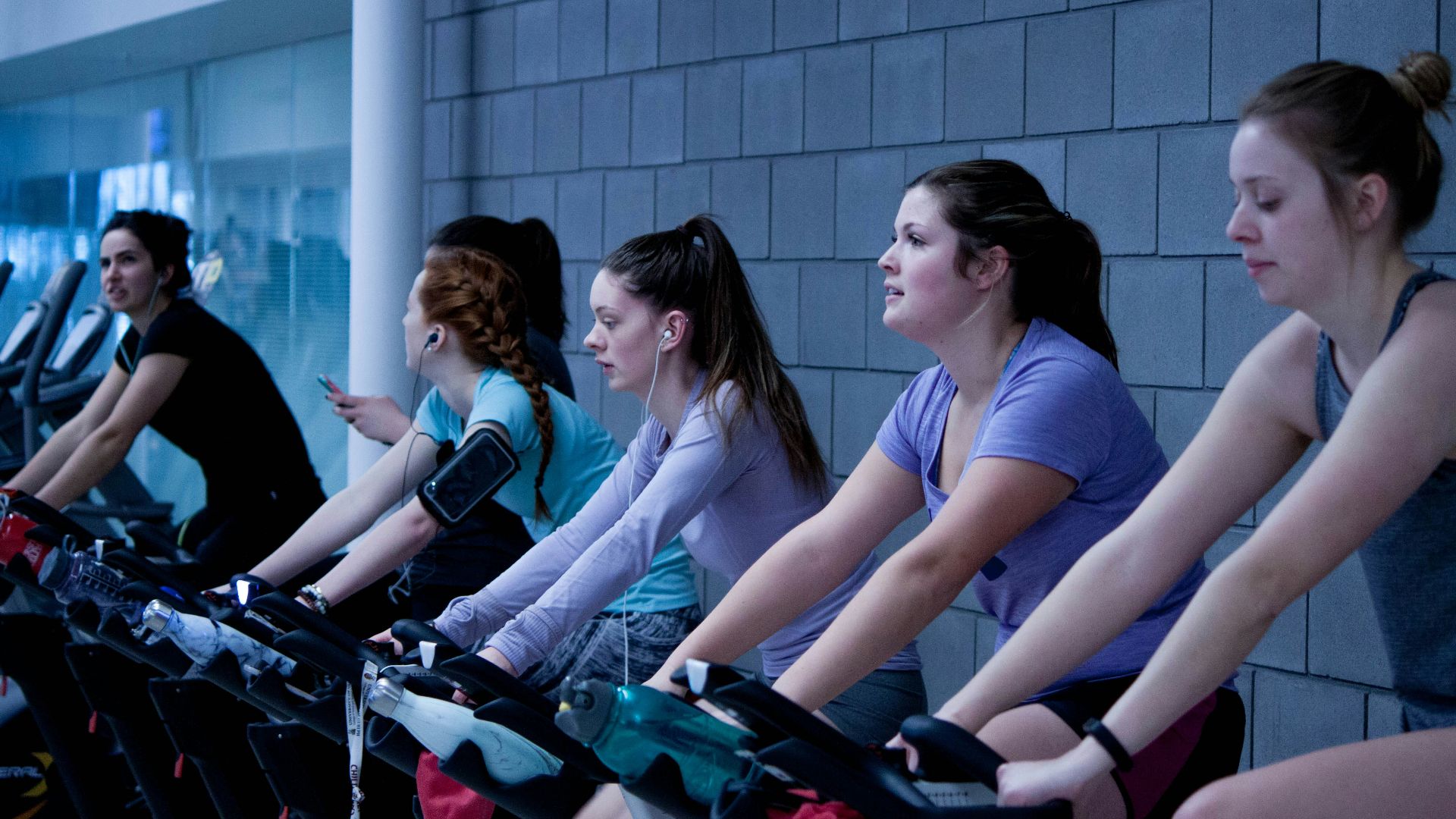 women taking exercise on black stationary bikes in front of gray concrete wall