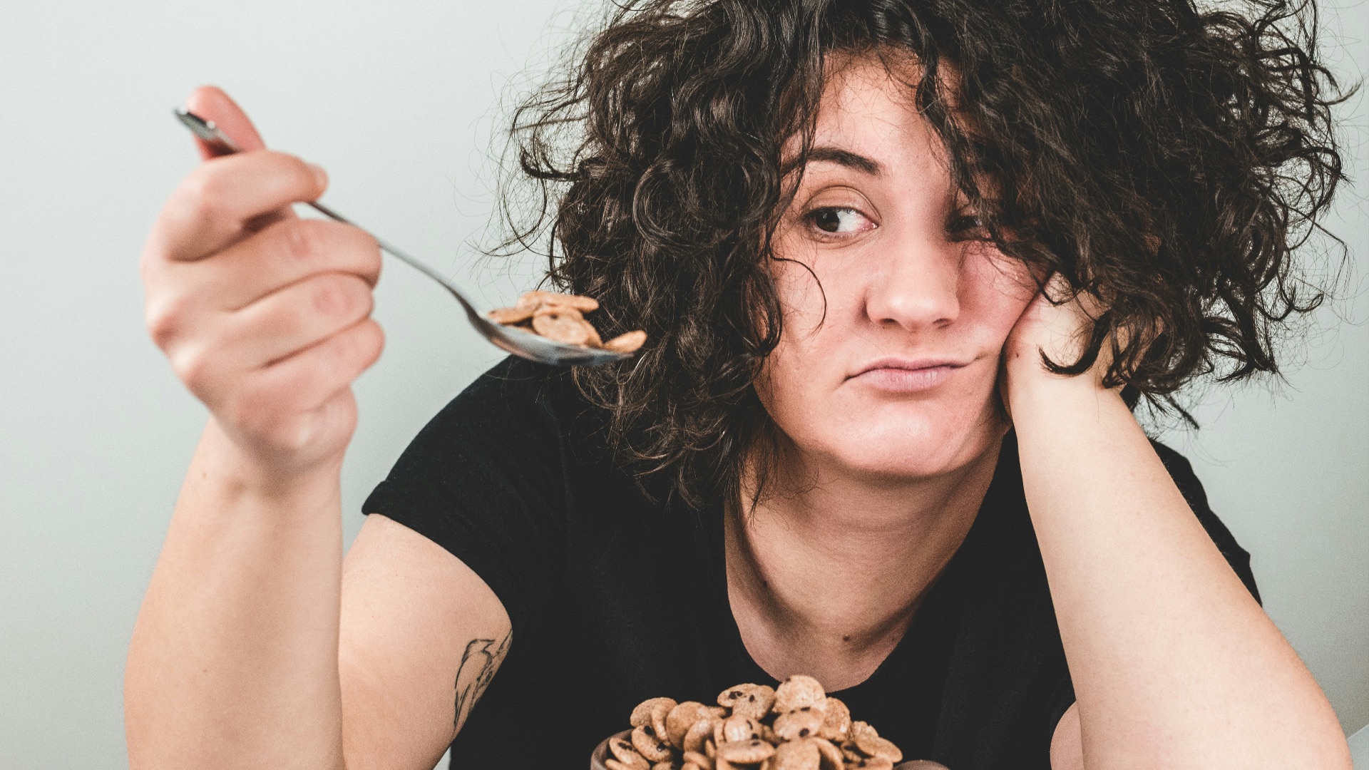 woman with messy hair wearing black crew-neck t-shirt holding spoon with cereals on top