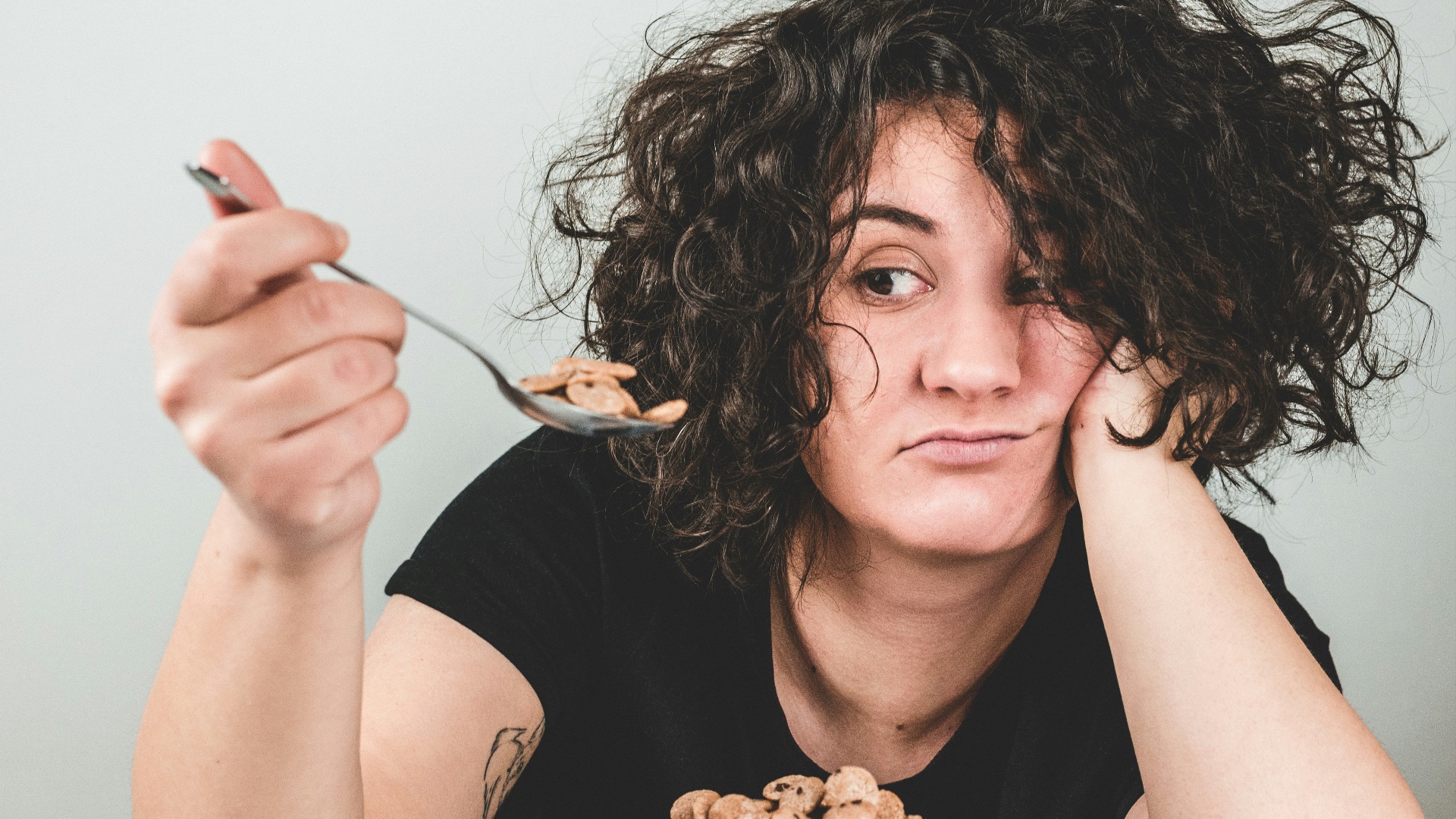 woman with messy hair wearing black crew-neck t-shirt holding spoon with cereals on top