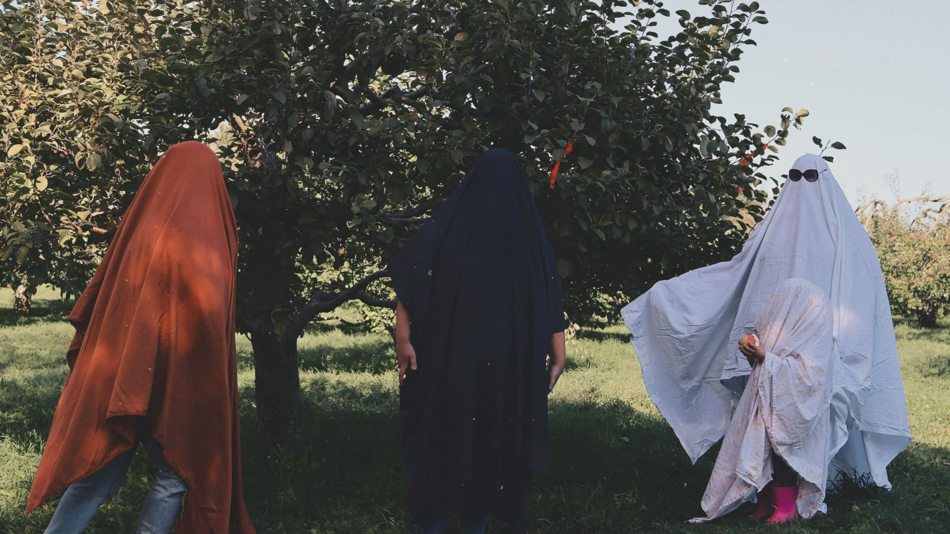 Three people in ghost costumes stand near a tree.