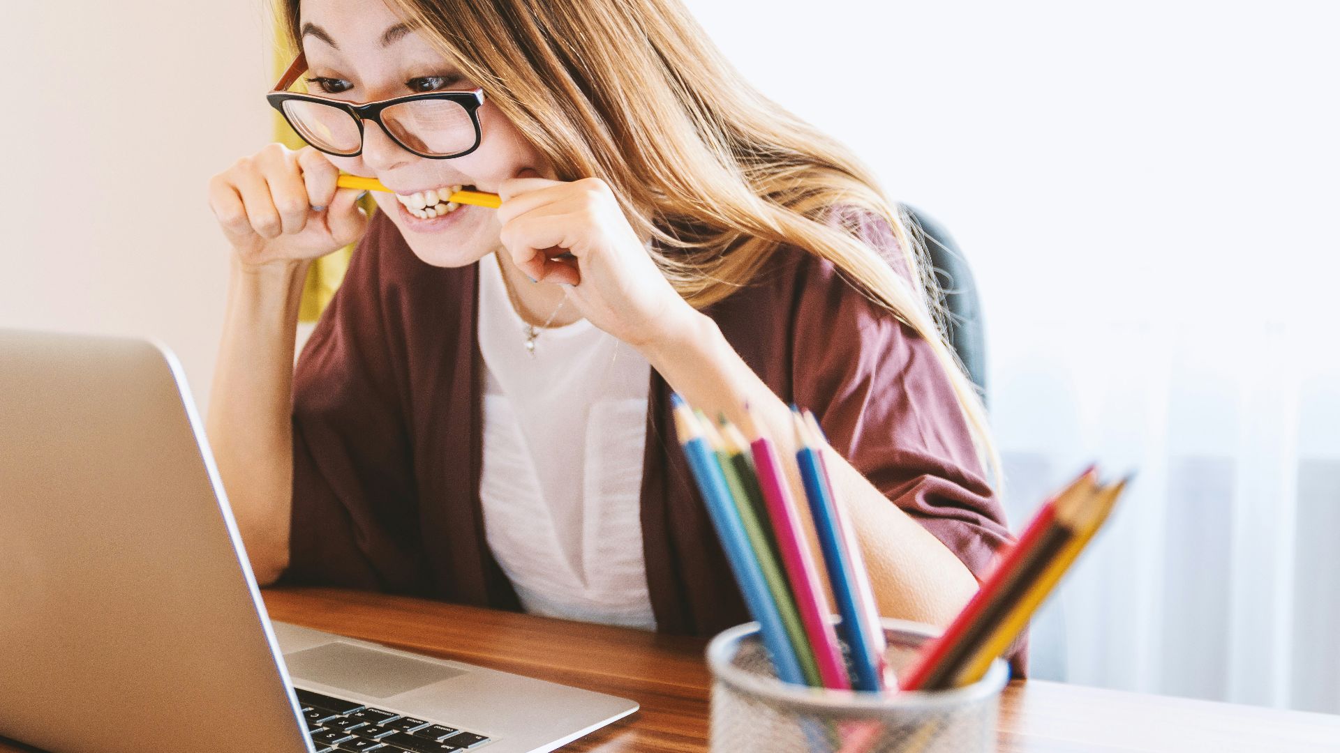 woman biting pencil while sitting on chair in front of computer during daytime