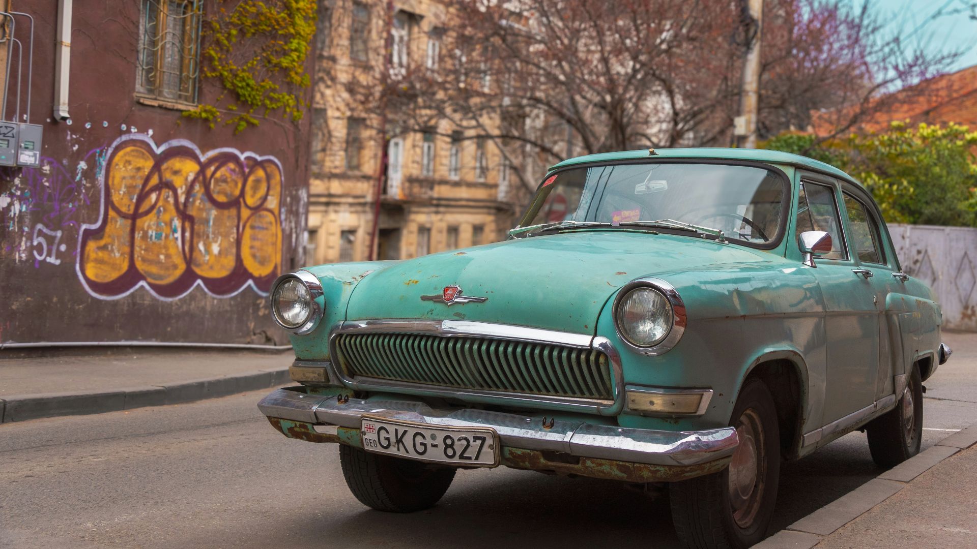 A green car driving down a street next to tall buildings