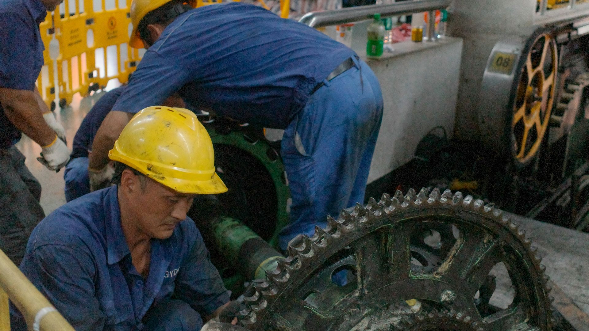 Workers in hard hats repair large industrial machinery gears.