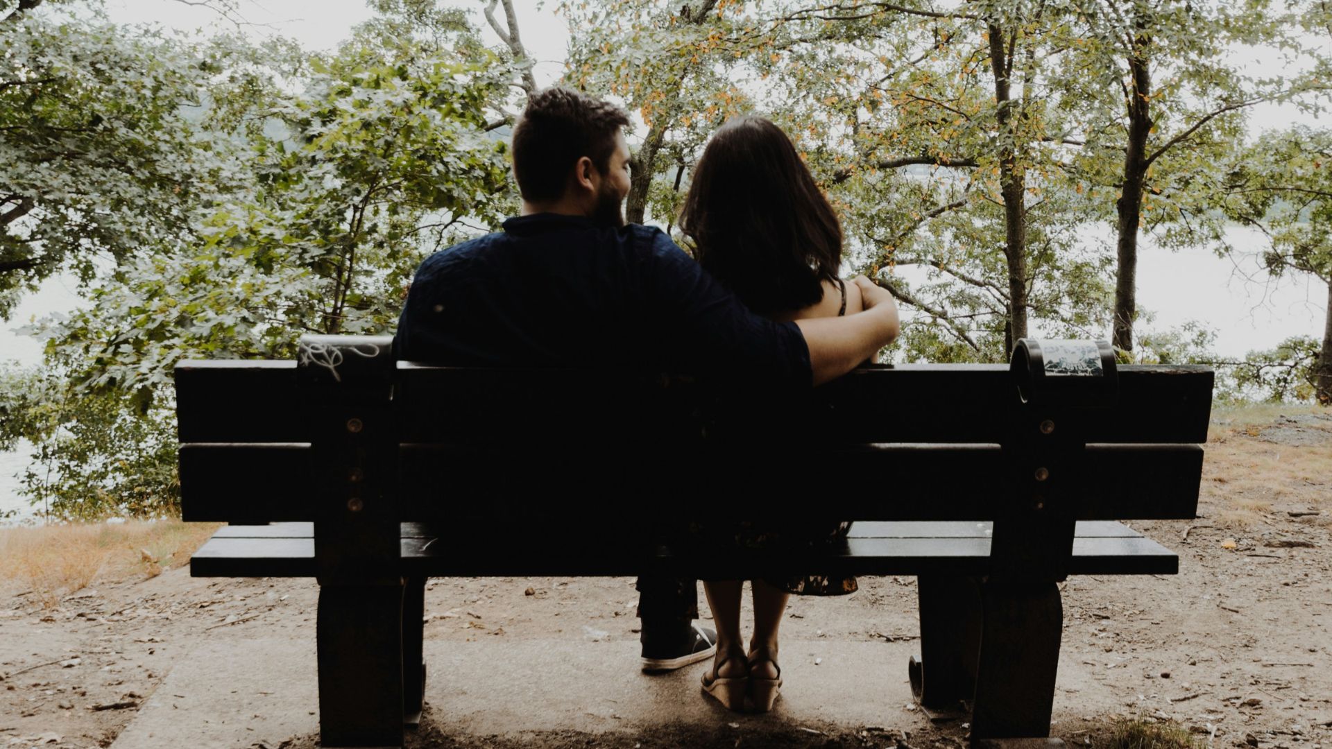 man looking to woman sitting on black wooden bench in front of tall trees during daytime