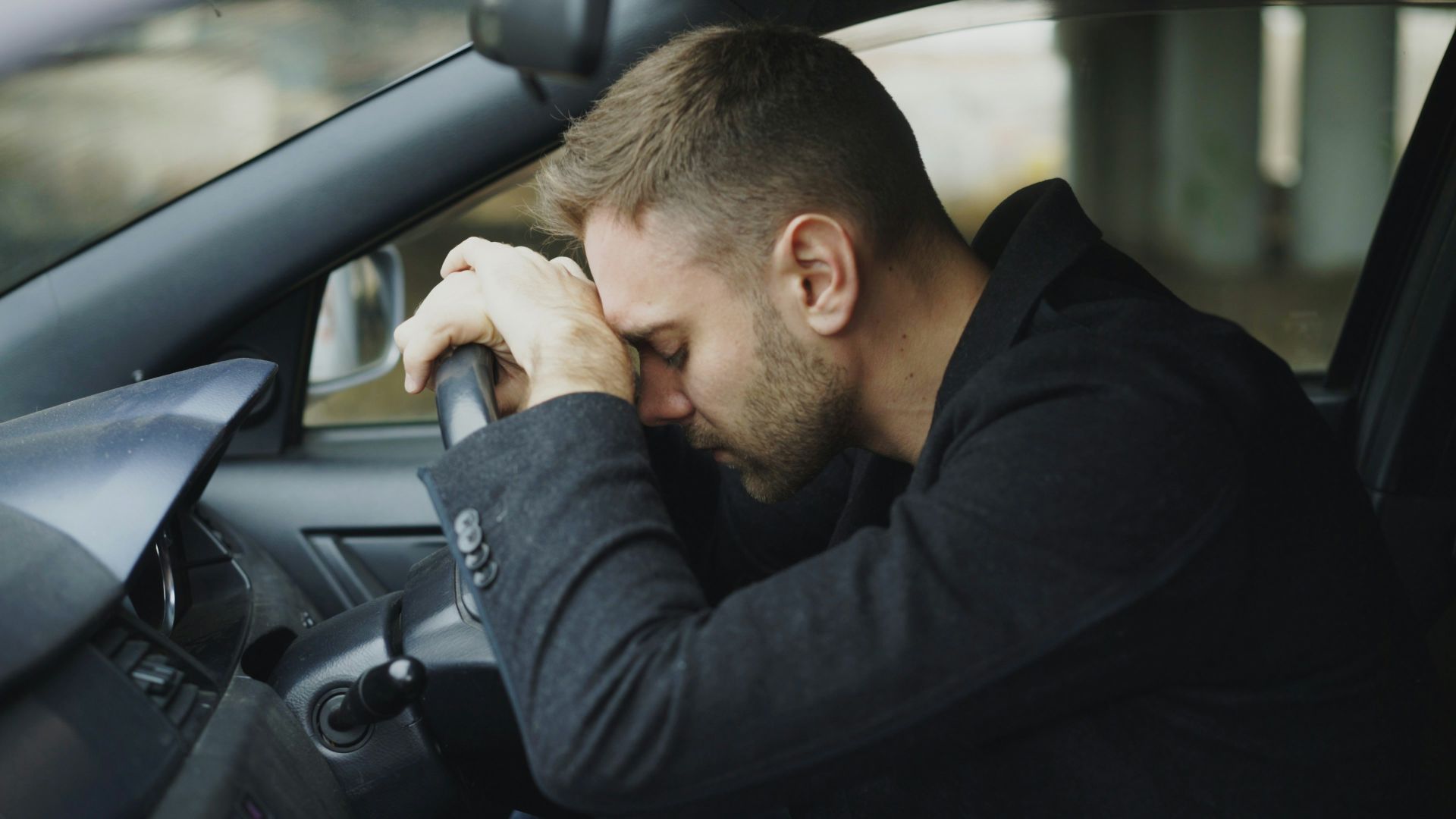 Man resting head on steering wheel in car.