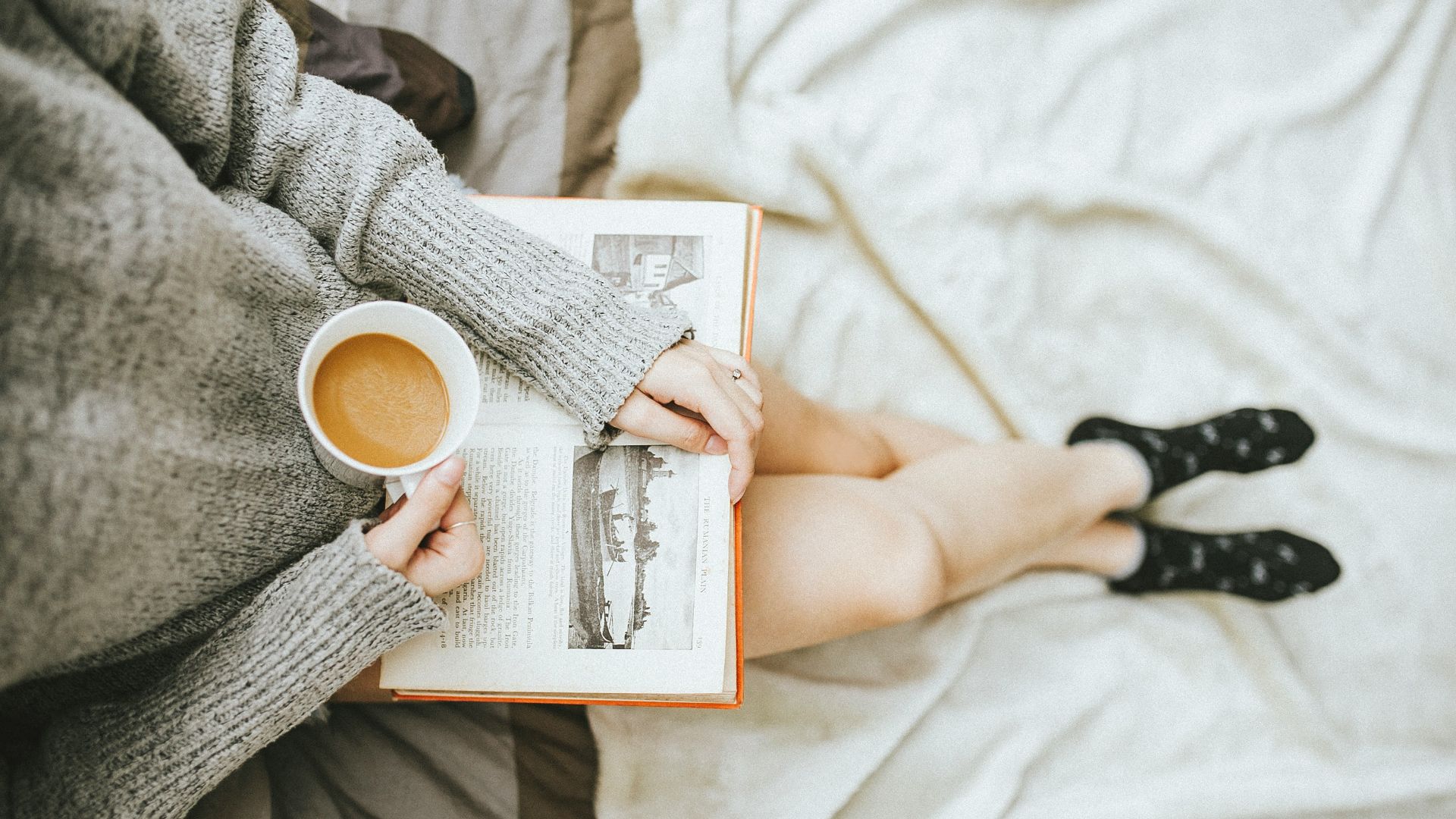 woman holding a cup of coffee at right hand and reading book on her lap while holding it open with her left hand in a well-lit room
