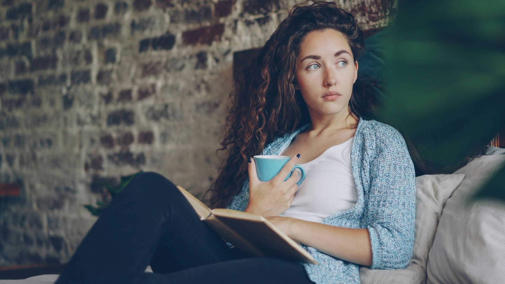 Woman holding coffee cup reading book on bed