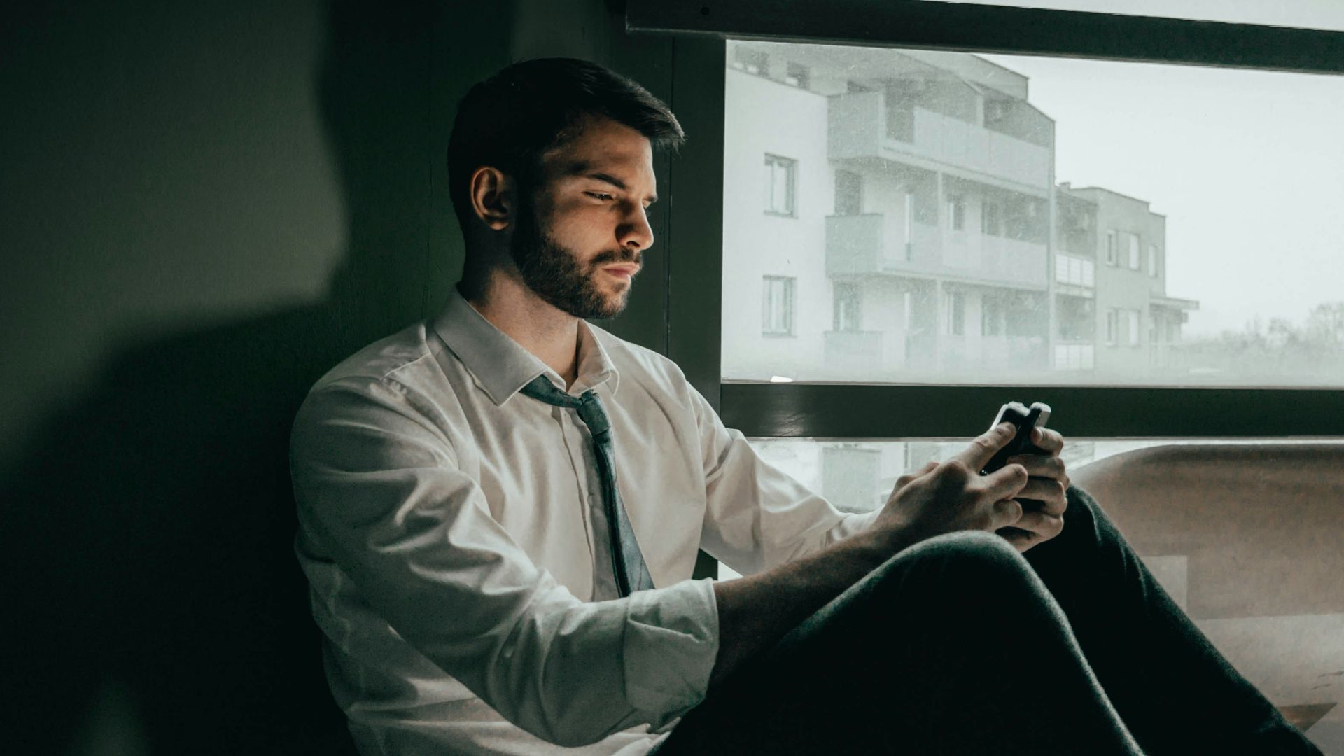 a man sitting on a window sill looking at his cell phone