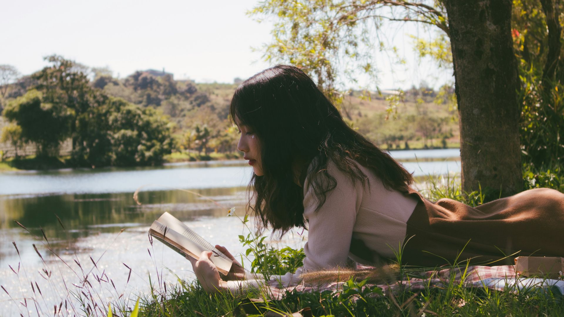 woman reading book and lying forward on sheet on grass beside body of water during day