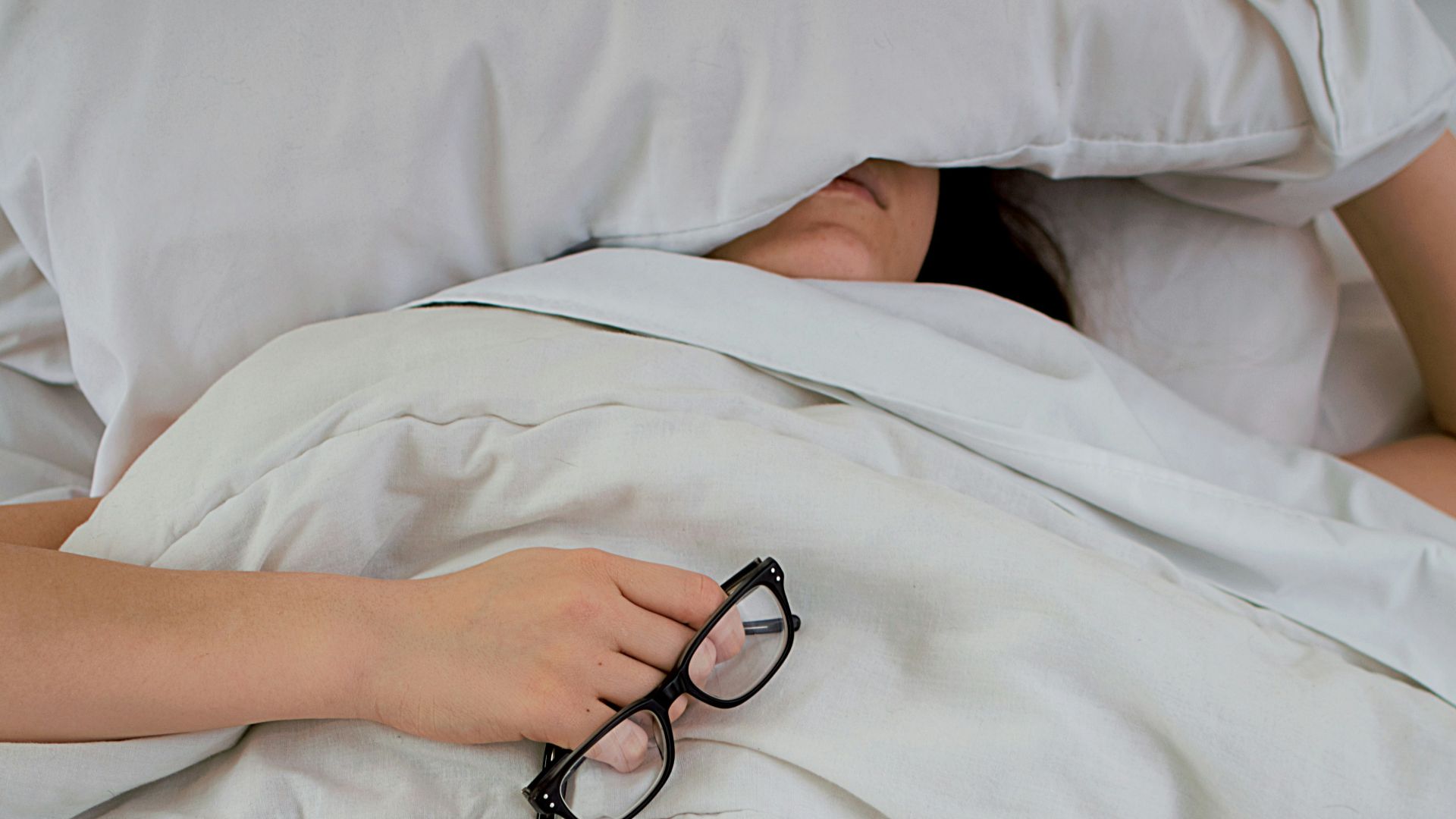 person lying on bed while covering face with pillow and holding eyeglasses