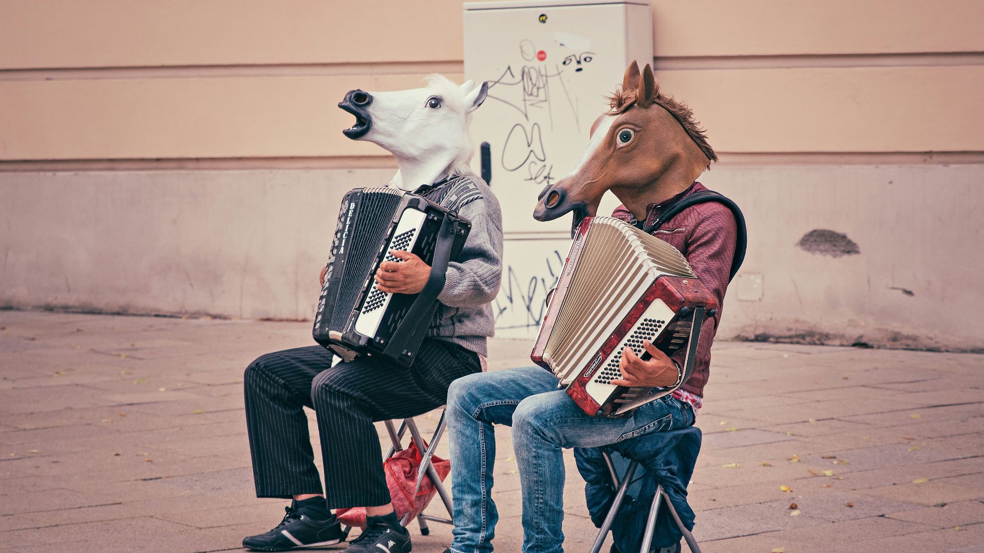 two person wearing horse heads sitting on folding chairs while playing accordions beside brown concrete building