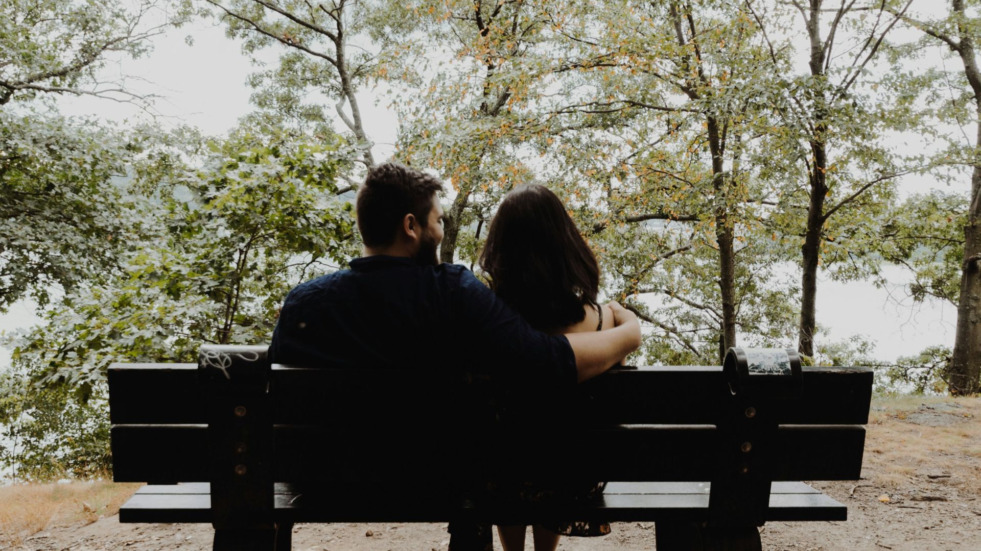 man looking to woman sitting on black wooden bench in front of tall trees during daytime