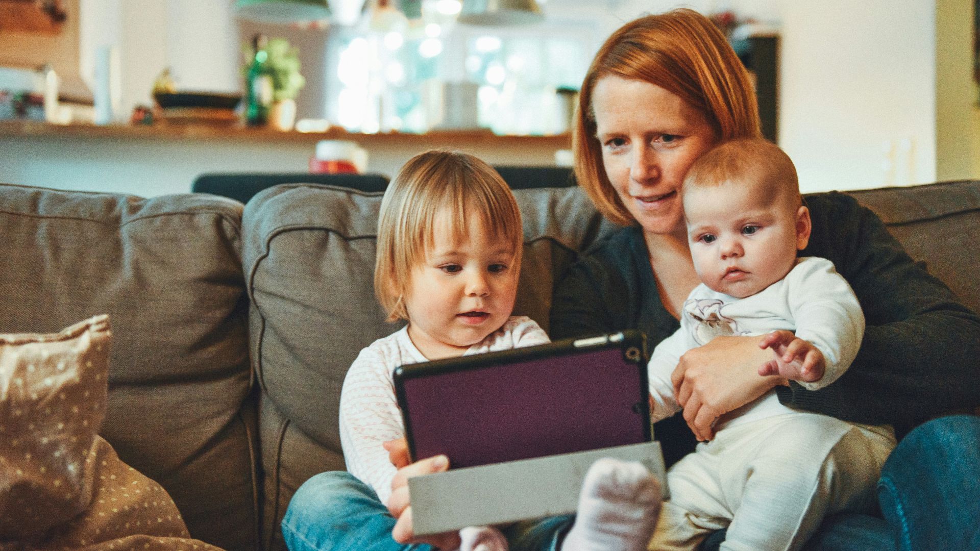 two babies and woman sitting on sofa while holding baby and watching on tablet