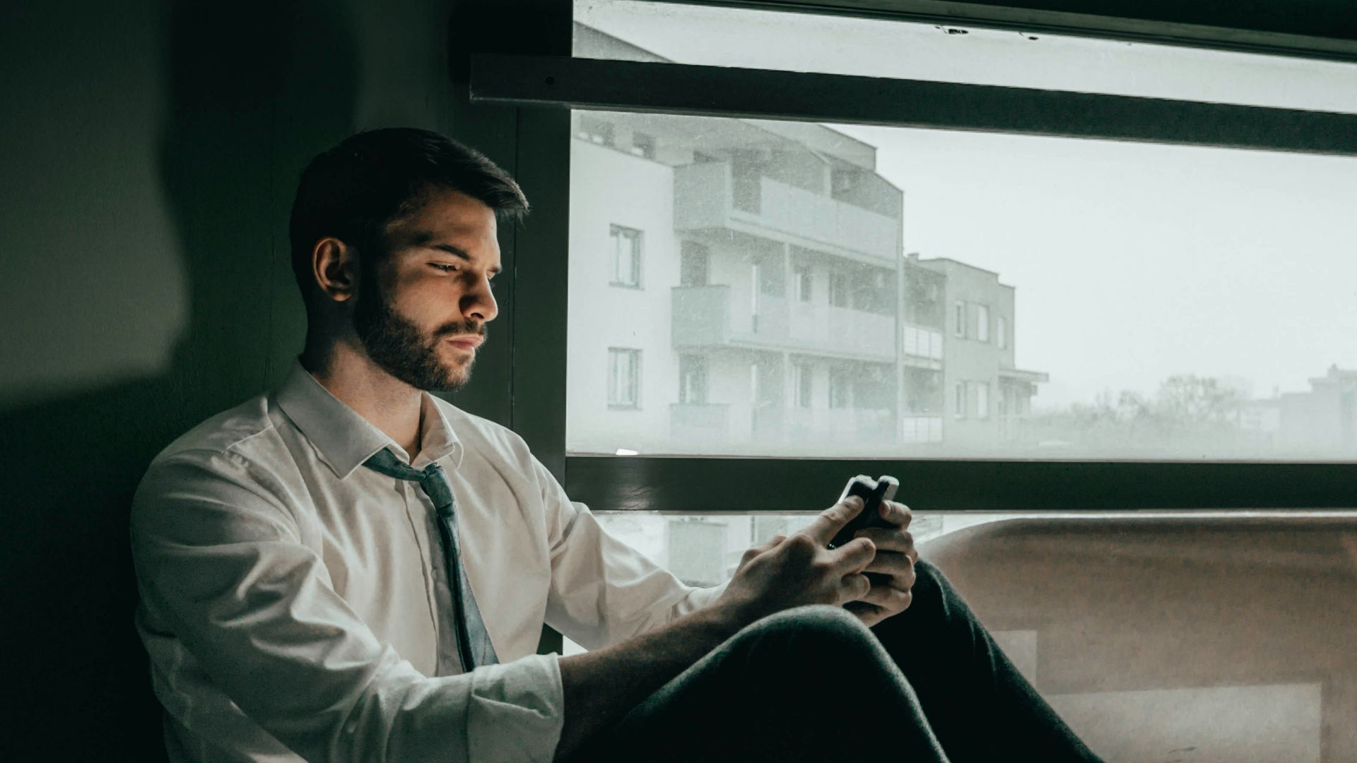 a man sitting on a window sill looking at his cell phone
