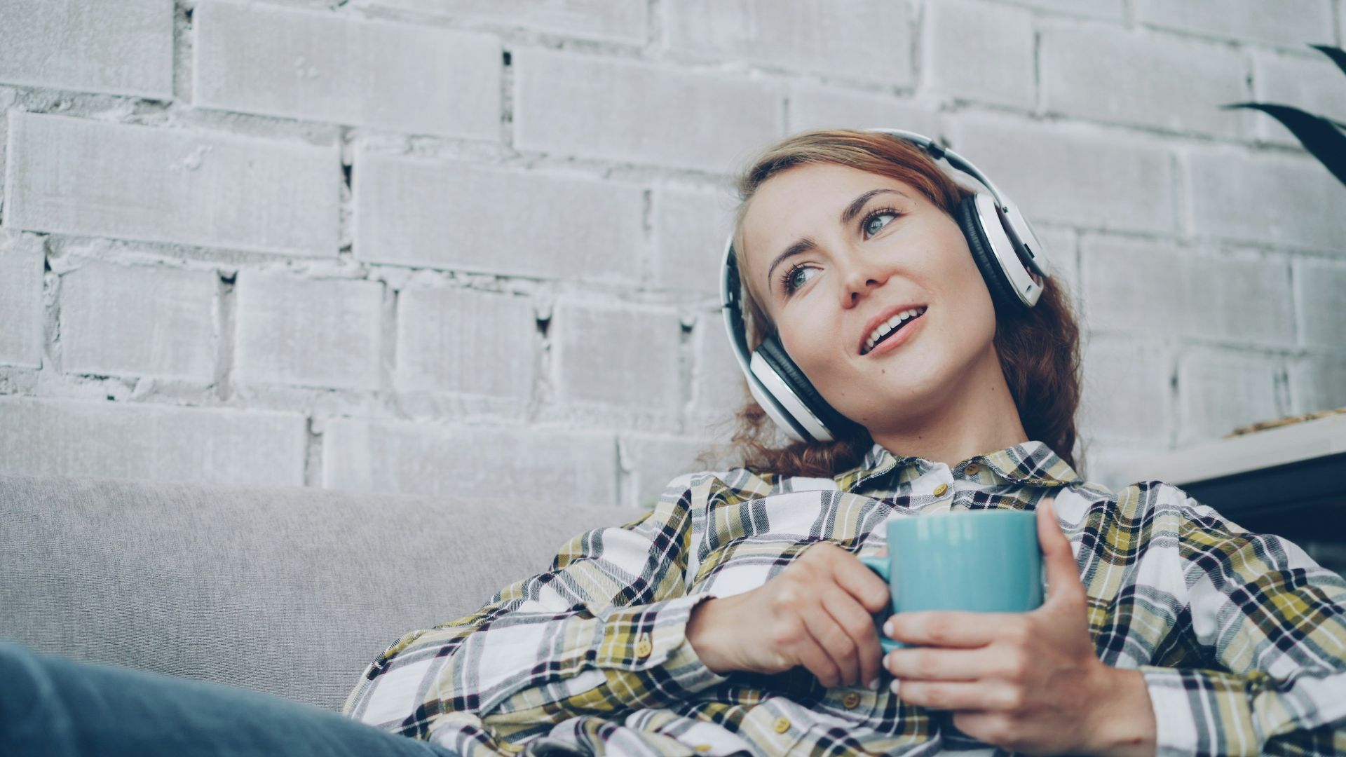 Woman wearing headphones holds a blue mug on couch