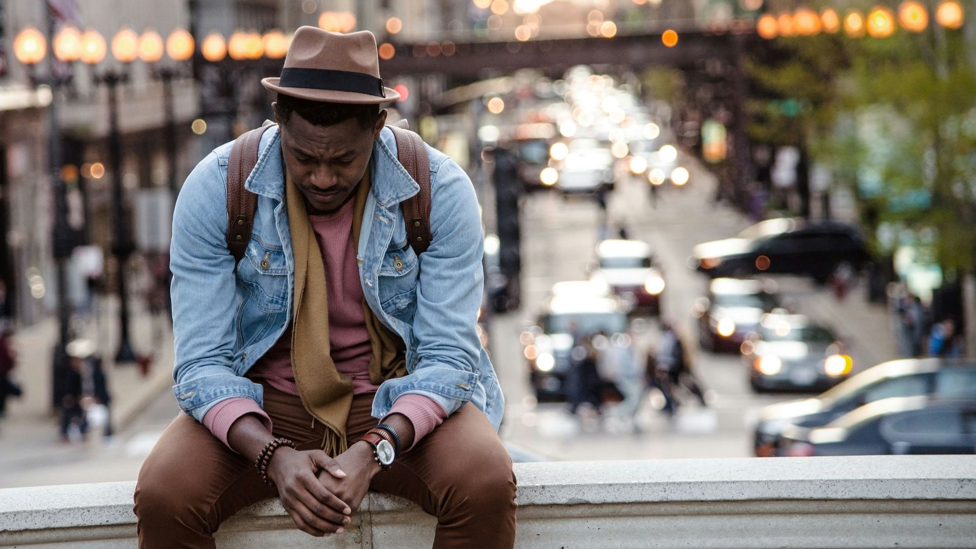 A man in a leather jacket looking down while sitting on a ledge in a city